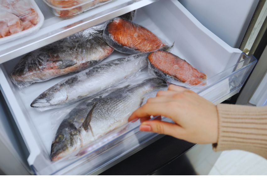 Mano sacando pescado congelado de un congelador, con varios filetes de salmón y peces enteros envueltos en plástico. Ideal para recetas de mariscos frescos.